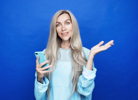 Foto de Modern lifestyle, old people, communication and electronic gadgets concept. Attractive elderly woman with long hair presents new smartphone over blue background. - Imagen libre de derechos