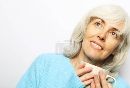 Téléchargez les photos : Une femme âgée aux cheveux gris porte un pull bleu avec un sourire mignon tenant une tasse de café sur fond blanc. Concept mode de vie, alimentation et personnes âgées. - en image libre de droit