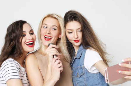 Téléchargez les photos : Three young female friends with a microphone singing and take picture with smartphone over white background - en image libre de droit