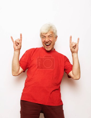 Téléchargez les photos : Vieil homme portant un t-shirt rouge décontracté debout sur un fond blanc isolé criant avec une expression folle faisant symbole de rock avec les mains en l'air. Une star de la musique. Concept lourd. - en image libre de droit