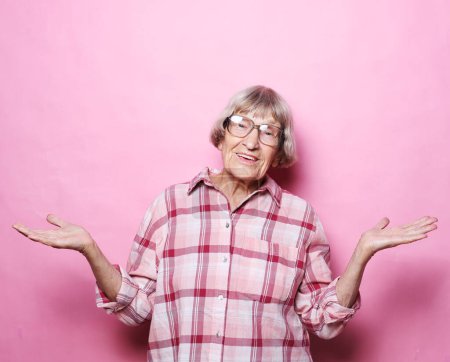 Téléchargez les photos : Portrait of an elderly woman with short gray hair, wearing eyeglasses, arms outstretched and smiling against a pink background.Old people, modern lifestyle, body language concept. - en image libre de droit