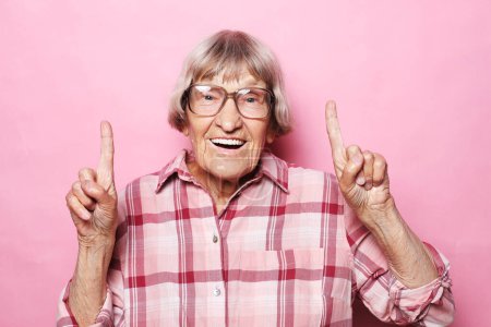 Téléchargez les photos : Portrait of a happy old woman wearing eyeglasses pointing upwards, isolated on a pink color background - en image libre de droit