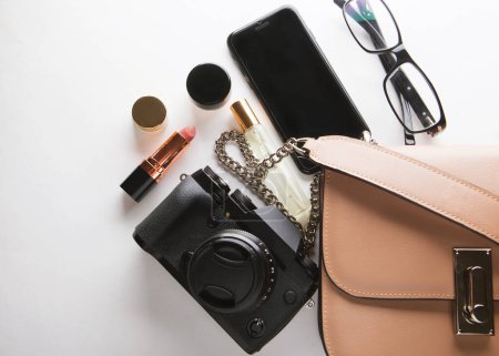 Foto de Bolso plano de mujer de cuero beige con cosméticos, accesorios, cámara y teléfono inteligente sobre fondo blanco
. - Imagen libre de derechos