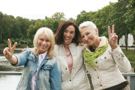Drei selbstbewusste Seniorinnen zeigen in einem Park in der Nähe des Sees V-Zeichen. Gruppe älterer Frauen in lässiger Kleidung. Reife Frauen genießen ihre goldenen Jahre.