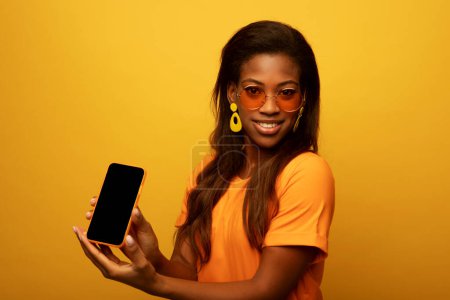 Téléchargez les photos : Young smiling african american woman isolated on yellow background, wearing orange t-shirt, holding blank phone and showing interesting offer over yellow background - en image libre de droit