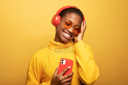 Téléchargez les photos : Image of beautiful african american woman in headphones singing and holding cellphone isolated over yellow background - en image libre de droit