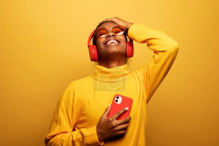 Téléchargez les photos : People, music, emotions concept. Delighted carefree afro american female with dances in rhythm of melody, closes eyes listens loud song in headphones, holds smart phone has fun. - en image libre de droit