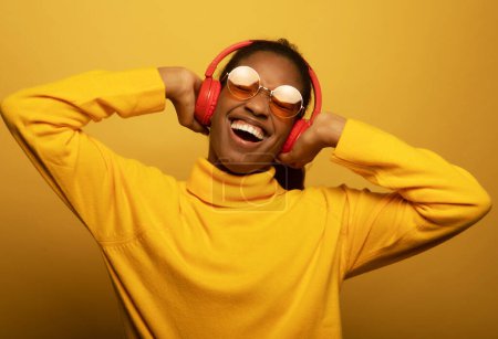 Foto de People, music, emotions concept. Young afro american female with dances in rhythm of melody, closes eyes listens loud song in headphones over yellow background. - Imagen libre de derechos
