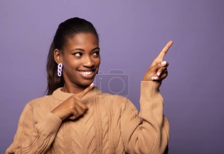 Téléchargez les photos : Happy african woman point finger right with excited cheerful smile. Young black female model present show new product or sale advertising space isolated over purple studio wall background - en image libre de droit