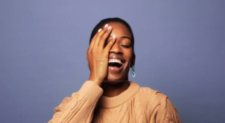 Téléchargez les photos : Portrait of a cheerful laughing happy young african woman who covers her eye with her palm and laughs posing against a blue background. - en image libre de droit