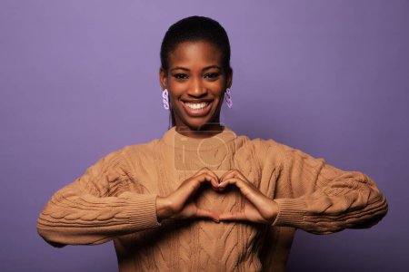 Téléchargez les photos : I love you. Portrait of thankful young african woman hold fingers joined in like sign. Headshot of loving black female looking at camera show heart symbol over violet background. - en image libre de droit