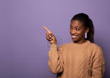 Foto de Happy african woman point finger left with excited cheerful smile. Young black female model present show new product or sale advertising space isolated over purple studio wall background - Imagen libre de derechos