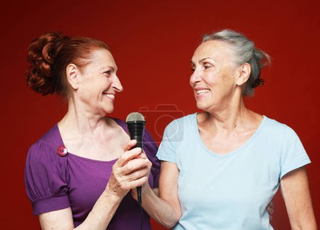 Foto de Lifestyle, party and old people concept: two elderly female friends with microphone, laughs and prepares for party karaoke over red background - Imagen libre de derechos