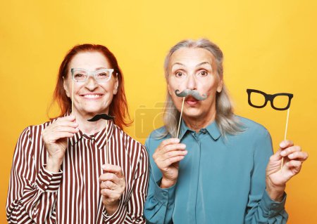 Foto de Lifestyle, party and old people concept: funny elderly female friends with fake mustache and glasses, laughs and prepares for party over yellow background - Imagen libre de derechos