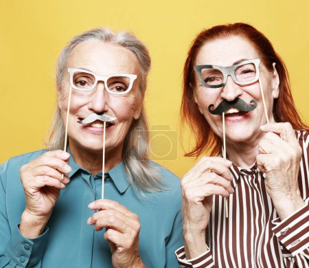 Téléchargez les photos : Lifestyle, party and old people concept: funny elderly female friends with fake mustache and glasses, laughs and prepares for party over yellow background - en image libre de droit