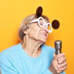 Emotion, modern lifestyle and old people concept: Happy old woman singing with microphone over yellow color background, having fun