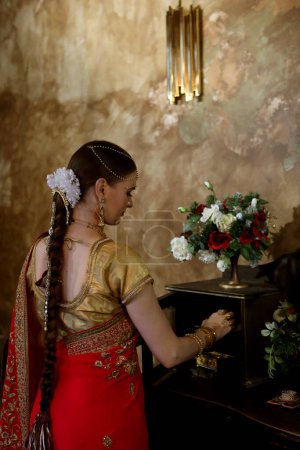 Téléchargez les photos : A young woman in a traditional Indian wedding dress opens drawers in a chest of drawers while standing with her back to the camera. Lifestyle concept. - en image libre de droit