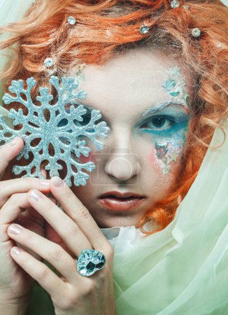 Foto de Party, christmas and people concept: A beautiful red-haired woman in a New Year's costume closes her eyes with a silver snowflake. Fairy, sorceress, fairy tale character. - Imagen libre de derechos