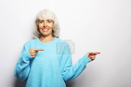 Téléchargez les photos : Femme âgée aux cheveux gris souriante portant un pull bleu isolé sur fond de studio gris pointant du doigt l'espace de copie vierge de côté, spectacle féminin heureux et positif à l'endroit publicitaire vacant - en image libre de droit
