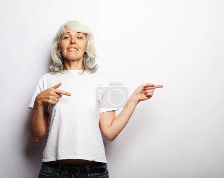 Téléchargez les photos : Femme aux cheveux gris âgée souriante isolée sur fond de studio gris pointant du doigt l'espace de copie vierge de côté, spectacle féminin âgé heureux positif à l'endroit publicitaire vacant - en image libre de droit