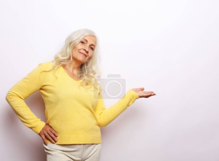 Foto de Sonríe mujer mayor con suéter amarillo sosteniendo algo en la palma abierta sobre fondo gris blanco - Imagen libre de derechos