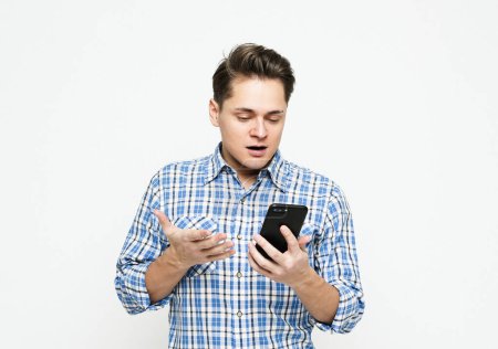 Foto de Portrait of a shocked young man wearing blue plaid shirt looking at mobile phone over white background - Imagen libre de derechos