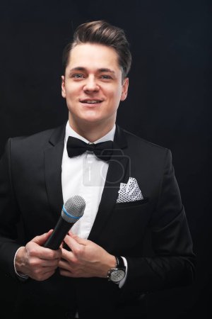 Photo for Stylish young man in a tuxedo holding a microphone, posing against a dark background with smoke, actor, singer, show, host of the event. - Royalty Free Image