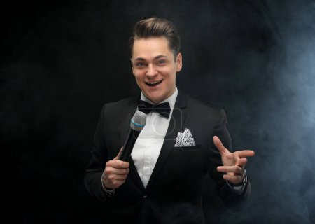 Foto de Stylish young blond man in a tuxedo holding a microphone, posing against a dark background with smoke - Imagen libre de derechos