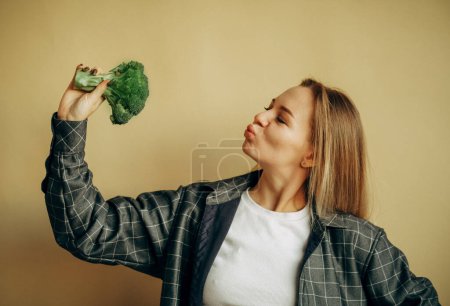 Photo for Photo of cheerful young blond woman standing over beige background while holding broccoli, close up - Royalty Free Image