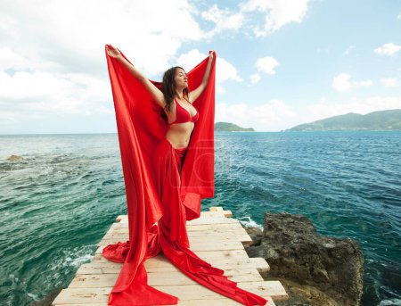 Foto de Young brunette woman with red skirt and fabric like a wings. female model near sea. Summer time. - Imagen libre de derechos