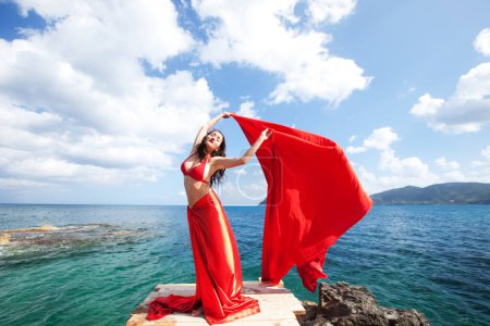 Foto de Young brunette woman with red skirt and fabric like a wings. female model near sea. Summer time. - Imagen libre de derechos