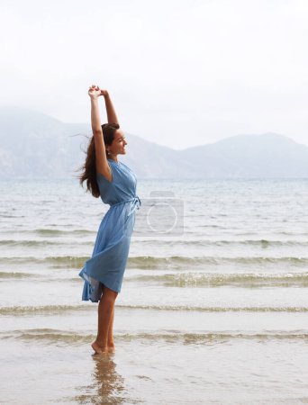 Foto de Mujer morena joven en un vestido azul caminando descalza en una playa y cuelga sus pies en el agua. Felices vacaciones de verano. - Imagen libre de derechos