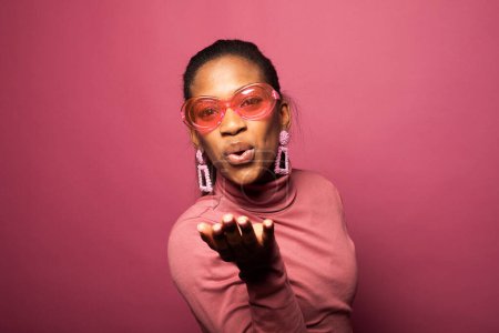 Foto de A cheerful afro american woman in a pink turtleneck blows a kiss to the camera. Portrait on a pink studio background. - Imagen libre de derechos