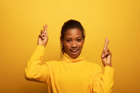 Foto de Attractive young afro american woman in yellow sweater smiling happily with fingers crossed making a wish. Concept of hope and dreams. Portrait on a yellow studio background. - Imagen libre de derechos