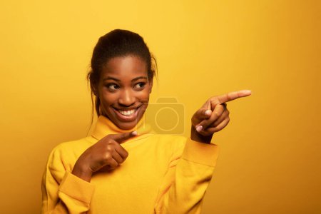 Téléchargez les photos : Cheerful african woman looking up and pointing finger to empty space for product, online courses or service advertising. Young black woman casual student gesturing showing place for ads - en image libre de droit