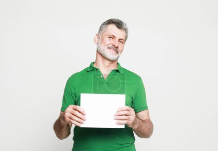 Foto de Senior handsome man with mustache and beard wearing green shirt holds the white sign in a studio over light grey background. Look here, great offer. - Imagen libre de derechos