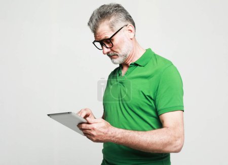 Photo for Photo of handsome mature man dressed green t-shirt and eyeglasses use tablet pc over light grey background. Tehnology and people concept. - Royalty Free Image