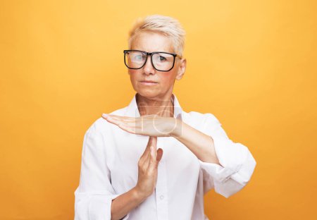 Foto de Gesture, prohibition and old people concept: Senior woman with short grey hair, wearing eyewear making time out sign over yellow background. - Imagen libre de derechos