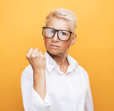 Foto de An elderly woman in glasses, with a short haircut, looks displeased and shows a clenched fist. Portrait on a yellow background. - Imagen libre de derechos