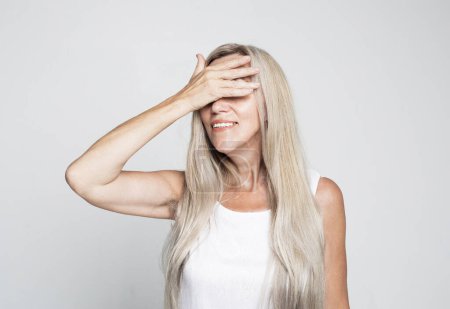Foto de Laughing mature woman 60 years old with long hair closes her eyes with her palm over grey background - Imagen libre de derechos
