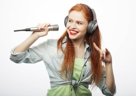 Photo for Cheerful young red-haired woman wearing headphones holding a microphone and singing. Portrait over white background. - Royalty Free Image