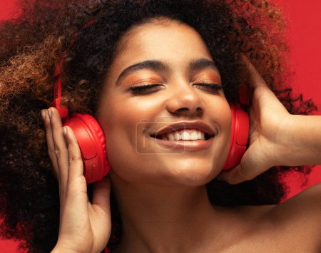 Photo for People, music, emotions concept. Young afro american female with dances in rhythm of melody, listens loud song in headphones over red background. - Royalty Free Image