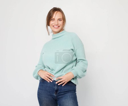 Photo for People, happiness and facial expressions concept. Pretty young blond woman wearing blue sweater with cute smile poses alone against light grey background. - Royalty Free Image