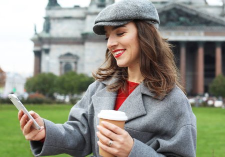 Foto de Retrato de una mujer joven usando su teléfono móvil mientras sostiene una taza de café en la calle. Concepto urbano y de comunicación. - Imagen libre de derechos