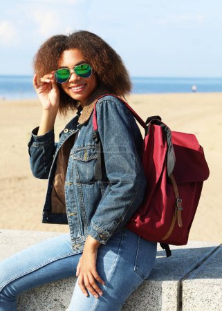 Téléchargez les photos : Beautiful young african woman in a denim jacket and sunglasses with backpack is resting while sitting on the beach on a summer day. - en image libre de droit