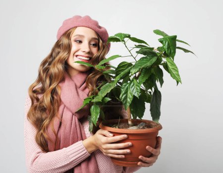 Photo for Attractive young woman with long wavy hair wearing pink hat holding a flower in pot over light grey background. - Royalty Free Image