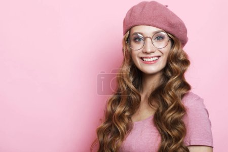 Foto de Beautiful young female model with long wavy hair wearing pink beret and eyeglasses. Portrait over pink color background. - Imagen libre de derechos