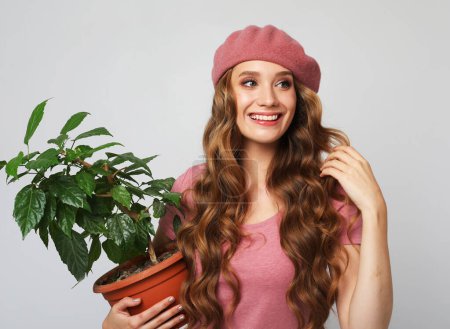 Foto de Attractive young woman with long wavy hair wearing pink hat holding a flower in pot over light grey background. - Imagen libre de derechos