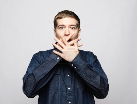Foto de A young handsome man covers his mouth with his palms over grey background. - Imagen libre de derechos