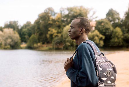 Foto de A young black man in sportswear with a backpack walks near the lake. Summer season. - Imagen libre de derechos